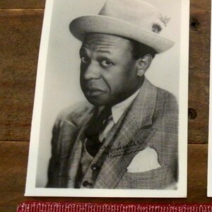 May include: A black and white vintage photograph of a person wearing a fedora hat and a plaid suit jacket with a pocket square. The photograph features the signature Rochester Eddie Anderson and is positioned above a red ruler measuring 4 inches.