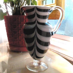 May include: A clear glass pitcher with a black and white striped design. The pitcher has a handle and a round base. The pitcher is approximately 9 inches tall.