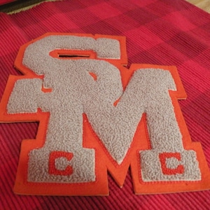 May include: A gray felt letter patch with the letters "SM" in orange felt. The patch is outlined in orange felt and has a small "C" in orange felt in the bottom corner of each letter.