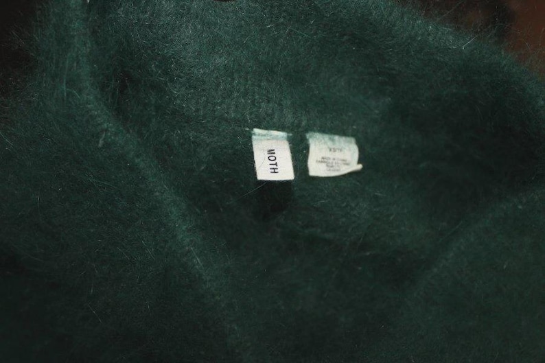 Op de afbeelding: Close-up van een donkergroene, fuzzy stof. Twee kleine witte labels zijn zichtbaar, &eacute;&eacute;n met de tekst "MOTH" en de andere met onleesbare tekst. De stof lijkt zacht en getextureerd, met een lichte glans.
