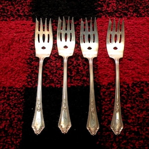 May include: Four silver forks with a face-like design on the tines, arranged on a red and black plaid background. The forks have a decorative pattern on the handles.