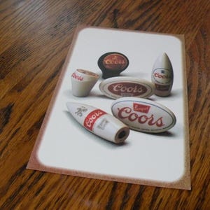 May include: A collection of six Coors beer tap handles. The handles are made of ceramic and feature the Coors logo in red and white. The handles are in various shapes, including a rocket, a bottle, and a circle.