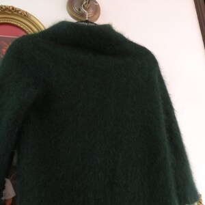 Op de afbeelding: Een donkergroene, donzige trui met een hoge kraag hangt aan een hanger. De textuur van de trui lijkt zacht en pluche. De achtergrond omvat een goudomrand schilderij en een witte muur.