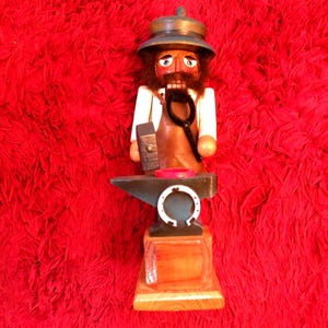 May include: A wooden nutcracker figurine depicting a blacksmith wearing a dark hat, white shirt, and brown apron. The figure holds a black hammer and tongs, standing behind a black anvil with a white horseshoe attached to the front base.