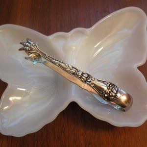 May include: A white, butterfly-shaped dish with a silver-colored sugar tong resting inside. The dish has a glossy finish, and the tongs are intricately decorated with floral patterns. The background is a dark wooden surface.