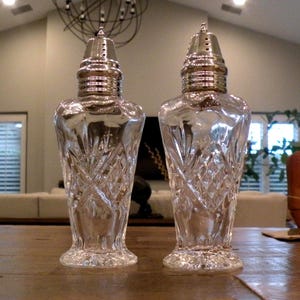 May include: A pair of vintage clear glass salt and pepper shakers with silver-colored metal tops. The glass shakers have a diamond-cut pattern and are displayed on a wooden surface. The shakers are approximately 6 inches tall.