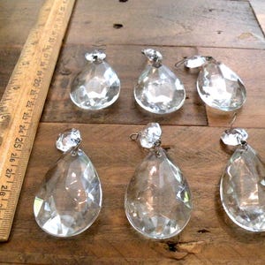 May include: Six clear crystal chandelier pendants with faceted surfaces, teardrop shape, and metal loops for hanging. A wooden ruler with inch markings is on the left side of the image.