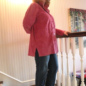 May include: A long-sleeved, button-down shirt in a deep rose-pink color with a textured finish, featuring a pointed collar, a straight hemline with side slits, and dark blue denim jeans worn underneath the shirt.