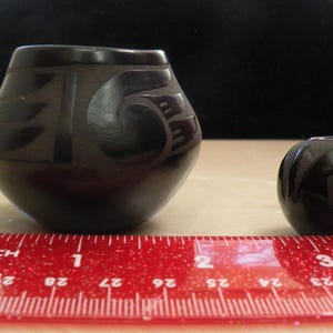 May include: Two black ceramic vessels with carved geometric patterns. The larger vessel is approximately 2 inches wide, and the smaller vessel is approximately 1 inch wide, as indicated by the red ruler placed in front of the objects.