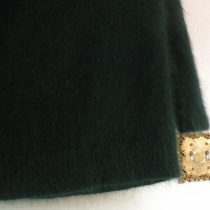 Op de afbeelding: Een donkergroene, donzige trui met een ronde hals en lange mouwen. De trui heeft een zacht, getextureerd uiterlijk. De afbeelding toont ook een goudkleurige lichtschakelaar.