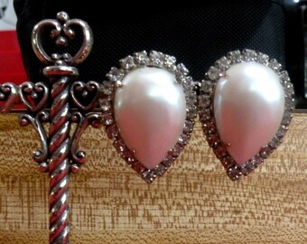 May include: A pair of teardrop-shaped white faux pearl earrings, each surrounded by a halo of small clear crystals. To the left, a silver-toned metal decorative element features a twisted vertical post and heart-shaped scrollwork details.