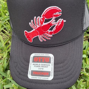 Op de afbeelding: Zwarte truckerpet met een rode kreeft geborduurd op de voorkant. De pet heeft een mesh achterkant en een druksluiting. De voorkant van de pet heeft een patch met de tekst "OTTO WORLD FAMOUS FOAM FRONT OSFM TRUCKER DURABLE SNAP CLOSURE".