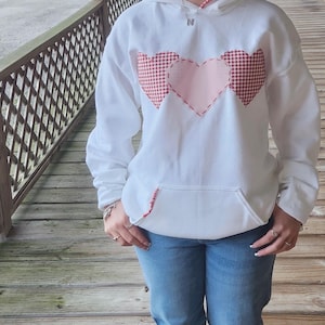 May include: White hoodie with a pink and red heart design. The hearts are made of fabric with a gingham pattern.
