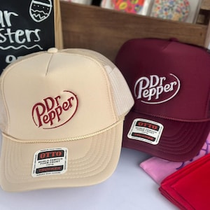 Puede incluir: Dos gorras de camionero con el logo de Dr. Pepper. Una gorra es de color beige con la parte trasera de malla beige, y la otra es de color granate. Ambas gorras tienen un logotipo blanco y cierre a presión.