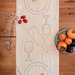Puede incluir: Un camino de mesa de lino blanco con bordado negro de dos platos de pasta, dos copas de vino y una botella de vino. El camino de mesa está sobre una mesa de madera con un plato de fruta y una rama de eucalipto.
