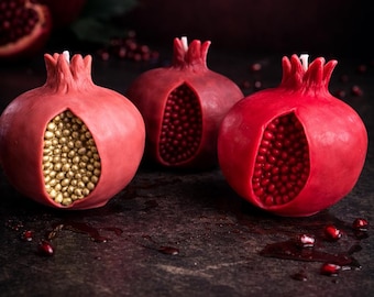 May include: Three pomegranate-shaped candles with a realistic design. One candle is open, revealing golden beads, while the others show red seeds. The candles are red with white accents and sit on a dark surface.