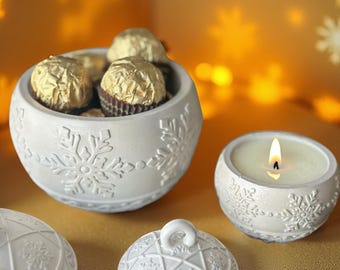 May include: White ceramic bowl filled with gold-wrapped chocolates, a lit candle, and a decorative lid. The bowl and candle holder feature snowflake and geometric patterns. The background is a warm, golden color.