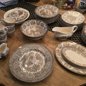 May include: A collection of vintage brown and white china tableware, including plates, bowls, cups, and serving dishes. The pieces display a detailed landscape pattern. The set is arranged on a wooden table, presenting a classic, elegant aesthetic.