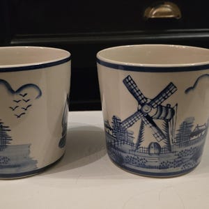 Op de afbeelding: Twee witte keramische mokken met blauwe en witte windmolenontwerpen. De mokken zijn versierd met een blauwe en witte windmolenscène, wolken en vogels.
