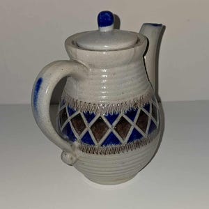 Op de afbeelding: Een kleine, grijze theepot van keramiek met een blauw en bruin ruitpatroon. De theepot heeft een deksel en een handvat.