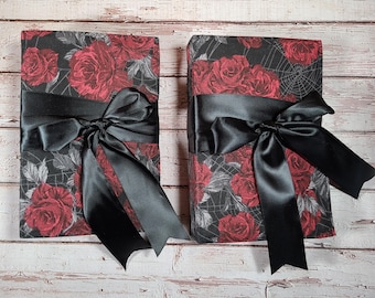 May include: Two black fabric-covered journals with a red rose and spiderweb pattern. Each journal is tied with a black satin ribbon.