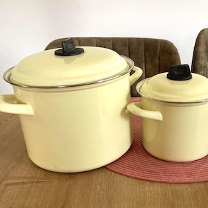 Op de afbeelding: Drie gele emaille pannen met deksels en handvatten. De pannen hebben verschillende maten, met een zilveren rand en zwarte dekselgreep. Ze staan op een rode mat op een houten tafel.