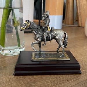 May include: A silver-coloured figurine of a soldier on horseback, mounted on a dark wooden base. The horse has gold-coloured accents on the bridle. The figurine is detailed and appears to be made of metal.
