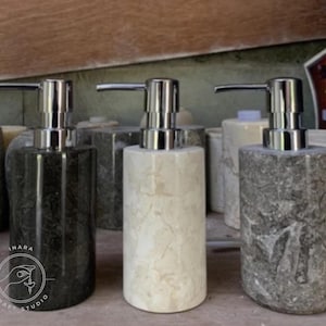 Op de afbeelding: Drie stenen zeepdispensers met zilveren pompkranen. De dispensers zijn in tinten zwart, crème en grijs, met natuurlijke aderingpatronen. De dispensers zijn cilindrisch en hebben een modern design.
