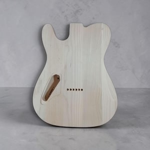 Pu&ograve; includere: Corpo di chitarra non rifinito in legno chiaro. Ha una forma Telecaster classica, con un singolo spacco e un bordo inferiore arrotondato. Include una cavit&agrave; di controllo e sei fori preforati per il ponte. La venatura del legno &egrave; visibile.