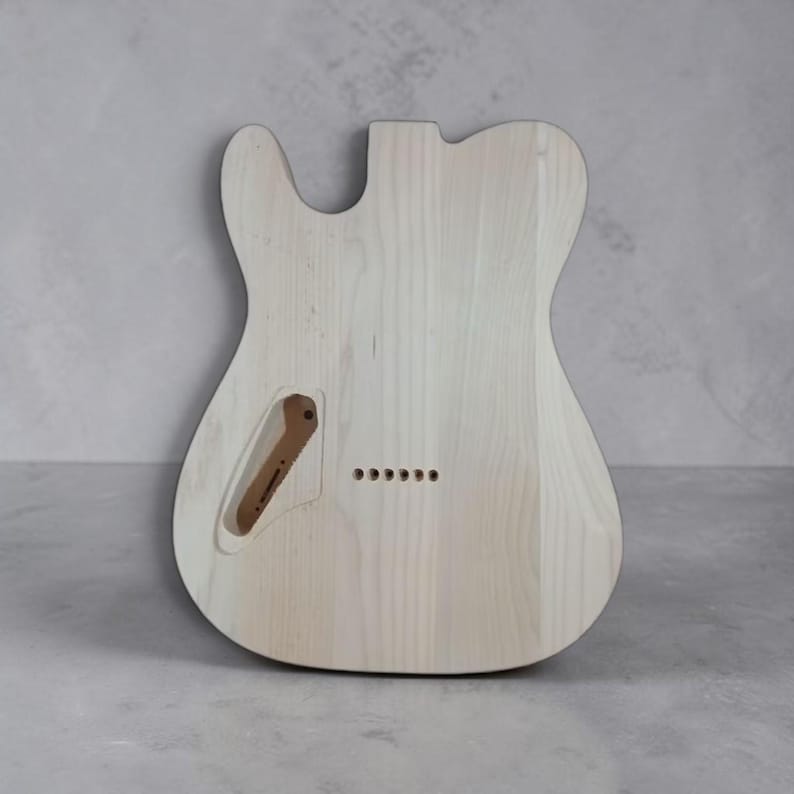 Pu&ograve; includere: Corpo di chitarra non rifinito in legno chiaro. Ha una forma Telecaster classica, con un singolo spacco e un bordo inferiore arrotondato. Include una cavit&agrave; di controllo e sei fori preforati per il ponte. La venatura del legno &egrave; visibile.
