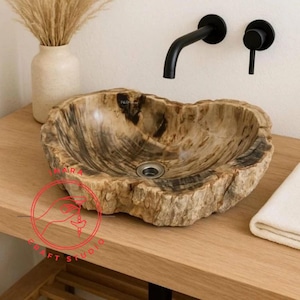 May include: A unique, petrified wood sink with a natural, organic shape. The sink features a central drain and is set on a wooden vanity. Black faucet fixtures are mounted on the wall above the sink. A vase with dried plants is on the left.