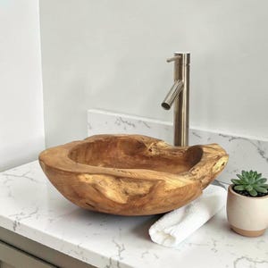 May include: A handcrafted wooden sink with a natural, organic shape. The sink is a warm brown color and sits on a white countertop. A brushed nickel faucet is installed. A small succulent in a pot and a white towel are also present.