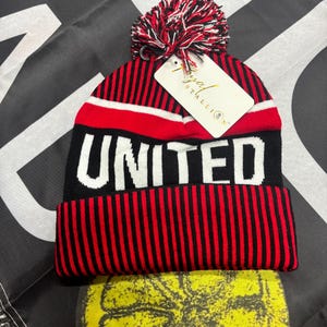 May include: A red, black, and white striped beanie with the word "UNITED" in large white letters. The beanie has a red, white, and black pom-pom on top and a tag that reads "Royal Stallion".