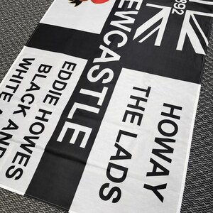 May include: A black and white flag with the text "Newcastle" and "Howay The Lads". Features a red poppy and the Union Jack flag. The flag also includes the text "Eddie Howes Black and White".