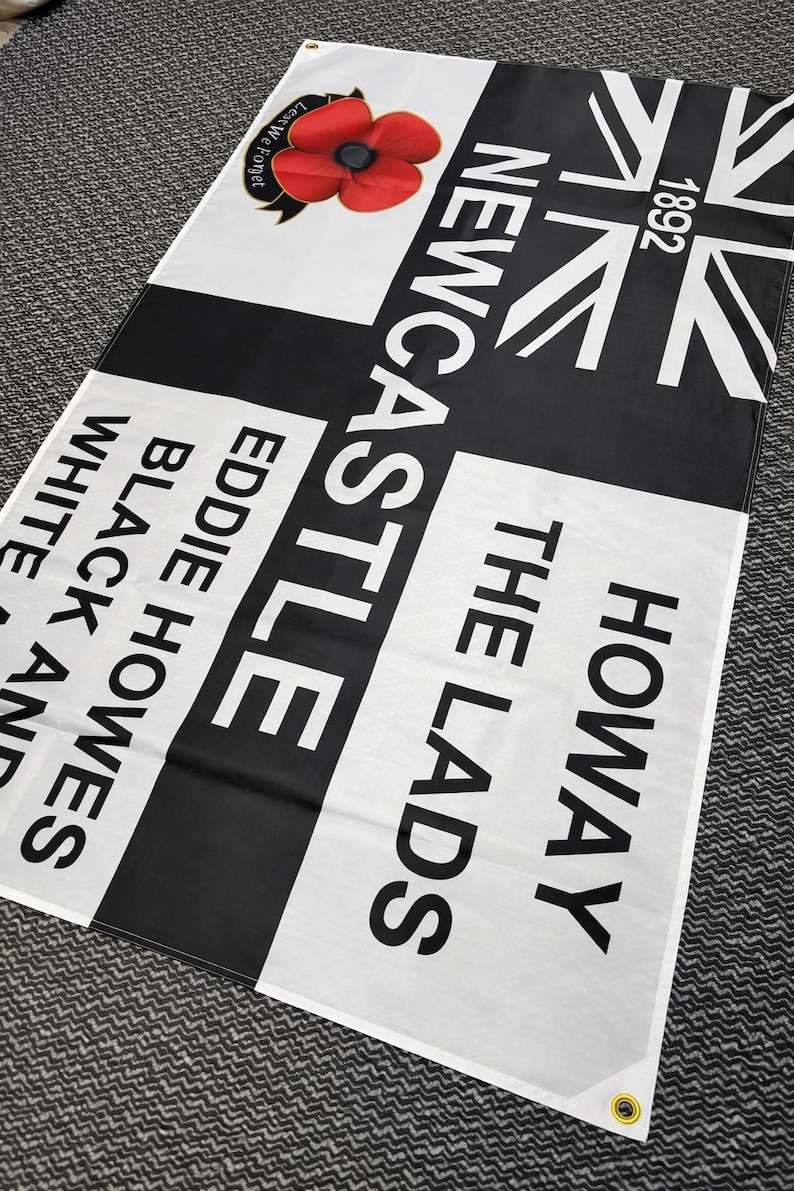 May include: A black and white flag with the text "Newcastle" and "Howay The Lads". Features a red poppy and the Union Jack flag. The flag also includes the text "Eddie Howes Black and White".