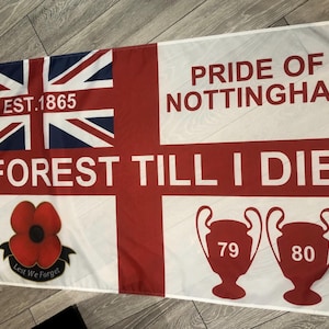 May include: A white flag with a red cross and a red poppy. The flag has the text "EST. 1865", "PRIDE OF NOTTINGHAM", "FOREST TILL I DIE", "79", and "80" on it. The flag also has the text "Lest We Forget" on it.