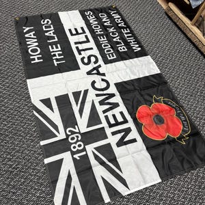 May include: A black and white flag with the text "HOWAY THE LADS" and "NEWCASTLE" in white. The flag features a Union Jack design and a red poppy with the words "Lest We Forget".