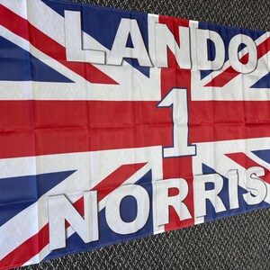 May include: A rectangular flag featuring the Union Jack design in red, white, and blue. The flag displays the name "LANDO NORRIS" in large white letters, with the number "1" in the center.