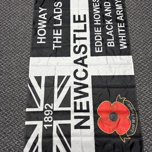 May include: Black and white flag with text reading "HOWAY THE LADS NEWCASTLE EDDIE HOWES BLACK AND WHITE ARMY" and a Union Jack design. Features a red poppy with "Lest We Forget" text.