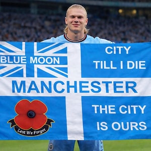 Puede incluir: Una bandera azul claro con detalles blancos y azules, sostenida por una persona. La bandera presenta un diseño de la Union Jack con "BLUE MOON" y "MANCHESTER". También tiene una amapola roja y las frases "CITY TILL I DIE" y "THE CITY IS OURS".
