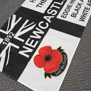 May include: A rectangular flag with a black and white design, featuring the word "Newcastle" in large white letters. The flag also includes the text "Howay The Lads," "Eddie Howies," and a red poppy with the words "Lest We Forget."