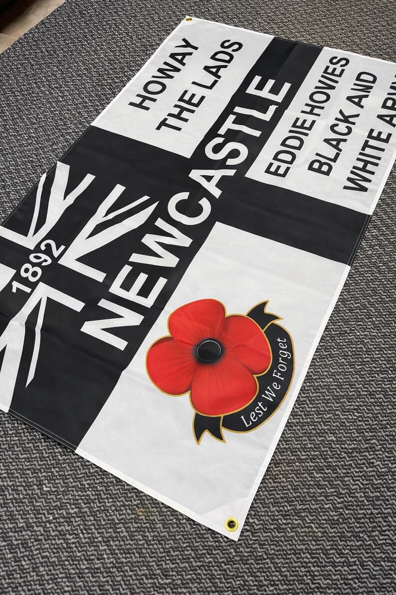 May include: A rectangular flag with a black and white design, featuring the word "Newcastle" in large white letters. The flag also includes the text "Howay The Lads," "Eddie Howies," and a red poppy with the words "Lest We Forget."