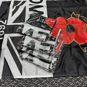 May include: Black and white flag with the word "Lancas" and a Union Jack design. Several folded flags are packaged in clear plastic. A red poppy flower is visible on the right side of the image.