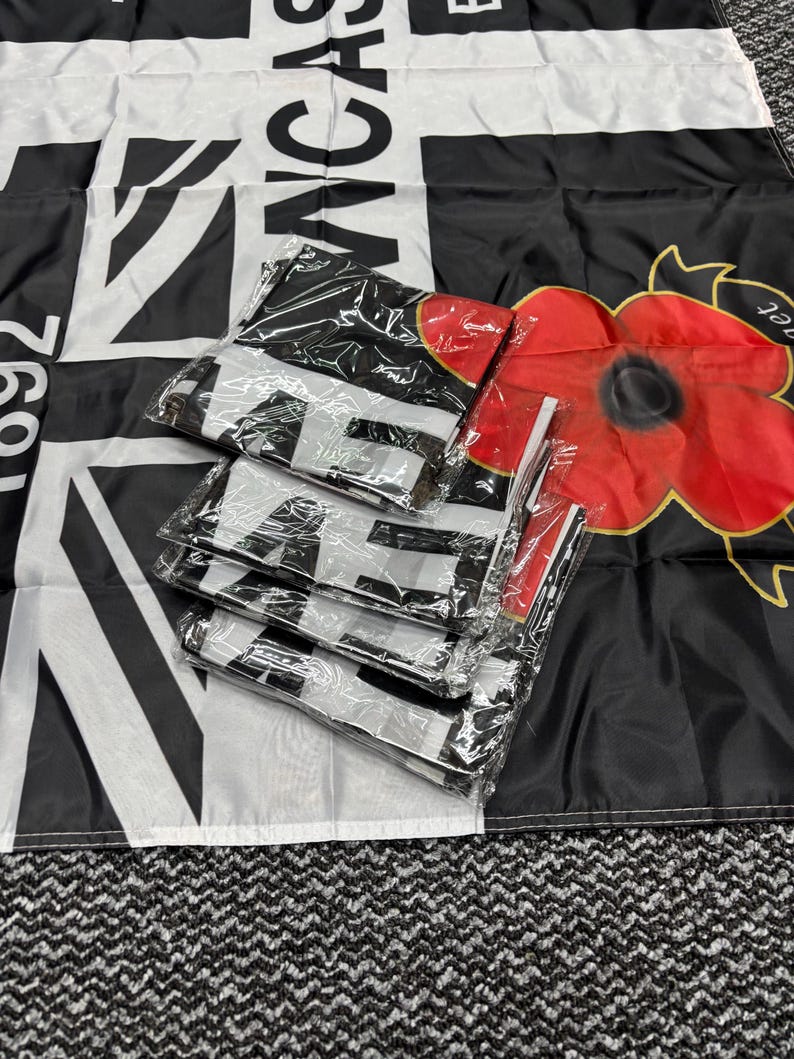 May include: Black and white flag with the word "Lancas" and a Union Jack design. Several folded flags are packaged in clear plastic. A red poppy flower is visible on the right side of the image.