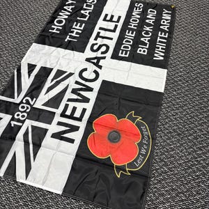 May include: Black and white flag with text that reads "HOWAY THE LADS" and "NEWCASTLE". Features a red poppy with the words "Lest We Forget". Includes the year "1892".