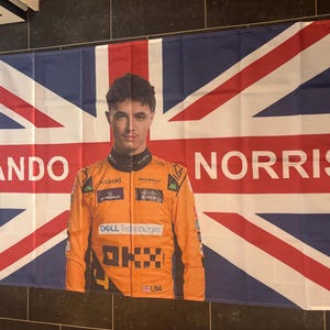Pode incluir: Uma grande bandeira com a Union Jack e um retrato de Lando Norris. A bandeira tem as palavras "LANDO" e "NORRIS" em letras brancas. Norris está usando um macacão de corrida laranja.