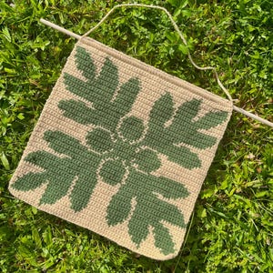 May include: A beige crocheted wall hanging with a green leaf design. The hanging is made with a simple stitch and has a rustic, natural look.