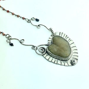 May include: A handcrafted silver necklace with a heart-shaped pendant. The pendant features a light green stone set in a textured silver frame. The necklace includes a delicate chain with pink beads and black accents, creating a unique, artistic design.