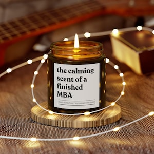 Puede incluir: Una vela de vidrio marrón con una etiqueta blanca que dice "the calming scent of a finished MBA". La vela está encendida y se encuentra sobre un posavasos de madera. La vela está rodeada de luces blancas de hadas.