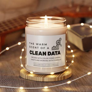May include: A lit soy candle in a glass jar with a white label that reads "THE WARM SCENT OF A CLEAN DATA." The candle sits on a small wooden coaster. The background includes a guitar and a book.